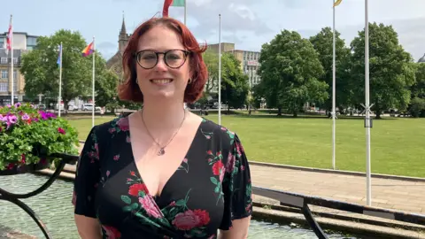 Sarah stands outside City Hall, smiling at the camera. She has short red hair and wears a black floral dress and round glasses. Behind her is a railing with a flower basket on it, part of a large water feature, several flagpoles and the green expanse of College Green. She is smiling at the camera and has her hands clasped behind her back.