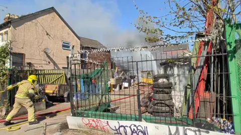 BBC A fire officer pulls a hose across an industrial yard containing tyres, trailers and scrap materials. Plumes of smoke and a jet of water are at the back of the yard.