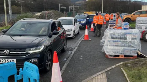 A team of workers is manning a water bottle station in Treorchy. The workers are wearing orange high-viz vests, and are stood around pallets of bottled water. It is next to a road where there are is a queue of people in cars waiting to be assisted. A large car is at the front of the queue with a window open.