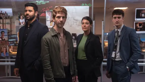 UKTV Three men and a woman police detectives, two wearing relaxed work attire and the other two in suit and tie, standing in front of a clear board with writing and images on them.