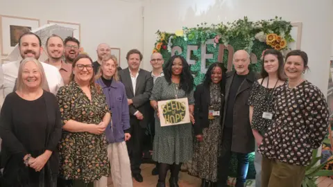 Calderdale Council About 15 people standing in a group. They are all looking and smiling at the camera. They are a mix of men and women. In the middle a woman holds a sign that says 'Seeds of Hope'