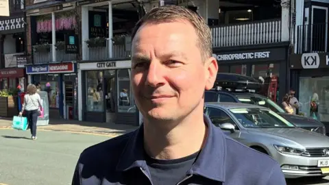 Carl Critchlow has short brown hair and is wearing a black t-shirt and blue jacket. He is standing next to a main road in Chester city centre near to shops. There are shoppers in the background.