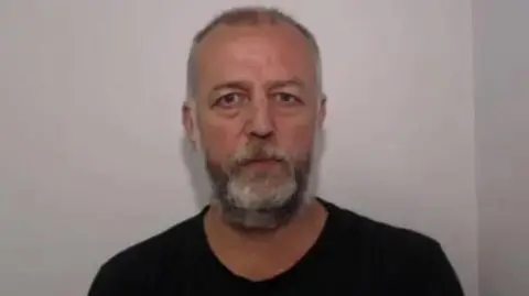 A custody image of Andrew Talbot wearing a black shirt in front of a grey called wall.