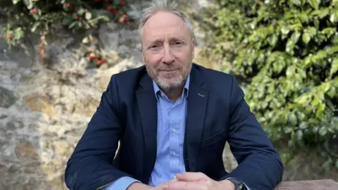 Deputy Paul Montague - A man with grey hair, a blue suit and a blue button up shirt. 