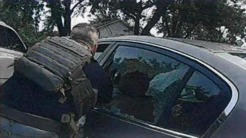 Person wearing tactical vest leans toward a car with shattered rear window, broken glass visible, trees and houses in background.