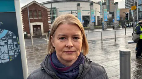 Anne Madden is standing in a street. It's been raining. Her fair hair is short. She's wearing a grey jacket and a blue and maroon scarf. The Waterfront Hall is behind her. 