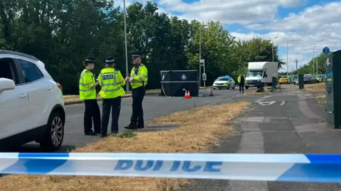 An incident behind a police tape cordon, with uniformed police officers talking and a black privacy screen erected in the middle of a road