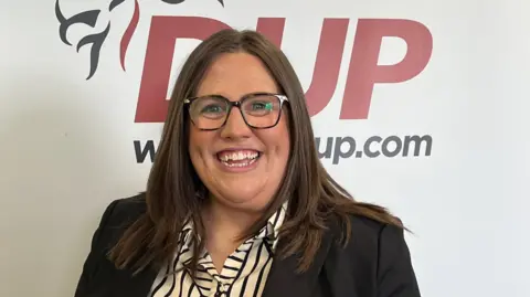 Shows a woman with glasses and smiling in front of a DUP banner, which says DUP and has a lion on the motif. 