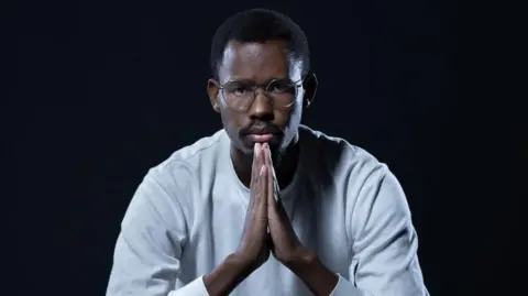 AFP Nelson Amenya, in a white top, holds his hands in front of him in prayer position while looking at the camera.