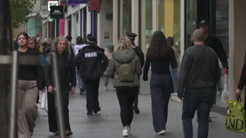 BBC Two police officers walking on a crowded Exeter High Street. There are lots of people walking both towards and away from the camera, some of them holding carrier bags and backpacks.
