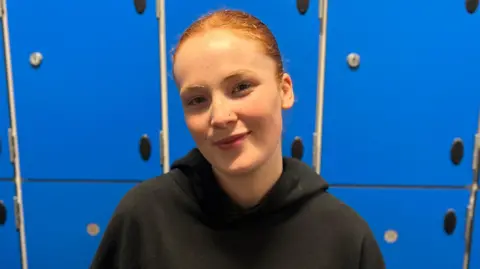 Jenna Crawford smiles at the camera, she is wearing a black jumper. She is standing in front of blue locker doors.