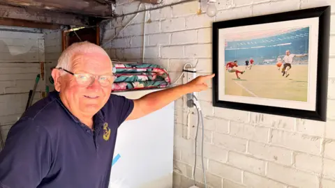 BBC A man in a navy blue T-shirt points at a picture of the 1966 World Cup game hung on a wall. The man is smiling.