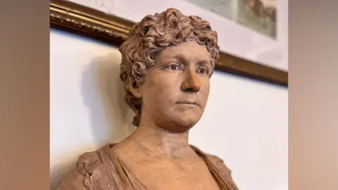 Theatre Royal Brighton A stone bust sculpture showing Ellen Nye Chart with short, curly hair, is set in front of a light wall with framed artwork above.