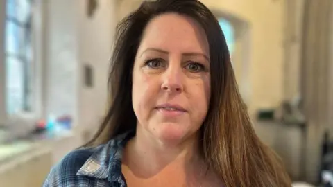Rachel Lockett is wearing a blue and white checked shirt. She has long dark hair and is looking at the camera.