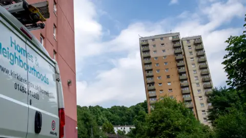 Kirklees Council A block of high-rise flats is in the distance, surrounded by trees. There is a white Kirklees Council van visible, with 'Homes and Neighbourhoods' written on the back doors in blue lettering.