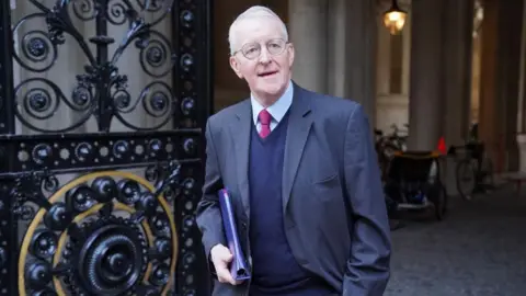 PA Media Benn is looking at the camera while walking out of a building. He is wearing a dark grey suit with a navy jumper underneath. He has a red tie on and light coloured shirt. He is holding a blue file under his arm.