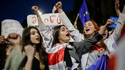 Three women gesture at a protest holding Georgian flags