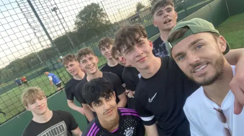 Mark James Maddison in a white shirt and green baseball cap, poses for a selfie with a group of teenagers wearing football kit.