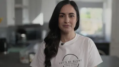 Leanne Lucas, who has long dark hair, smiles at the camera while wearing a white t-shirt with the logo 'Let's Be Blunt - Remove the Point'