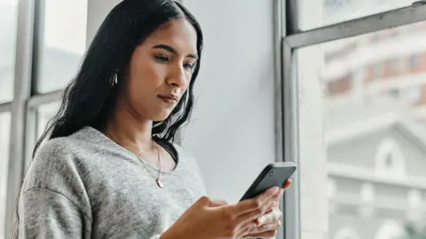 Getty Images Young woman in grey pullover standing by a window and looking at her phone with a serious look on her face.