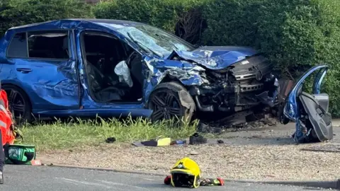 A side view of a crumpled up blue car on a pavement and half buried in a thick hedge. Its bonnet has buckled, its windscreen is a haze of cracks and its driver's side door has been removed and is resting on a driveway in front of the bonnet. There is a yellow emergency services helmet resting on the road in front of it, beside some gloves.