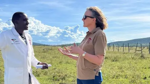 Harriet Gordon-Brown Harriet Gordon-Brown is standing next to a man in a lab coat, gesturing with her hands. They are out in a field with mountains in the background.