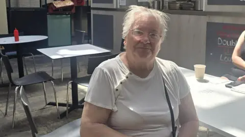 Hayley Coyle/BBC A person wearing a white short-sleeved top and glasses is seated at a white table in a cafe or food court setting. Several empty black chairs and tables are visible nearby, along with a paper cup on the table. In the background, a service counter and menu signage can be seen, with sunlight creating bright patches and shadows across the seating area.