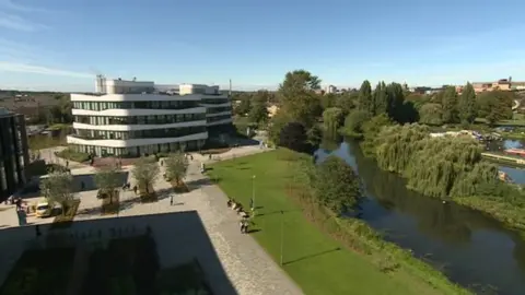 Aerial shot of University campus, showing four-storey teaching building in the background, river to the right with trees on the bank, grass area towards the centre and a row of small trees to the left.