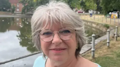 A woman with grey hair and glasses and wearing a baby blue jumper standing in front of a partially-full Crammer pond in Devizes, with houses and trees in the background. 