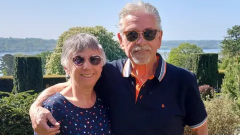 Woman and man, both wearing sunglasses, smile looking into the camera. The man is taller than the woman and has his arm around her. There are trees and greenery behind them.