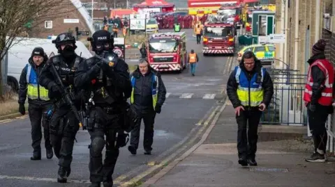 Northamptonshire Police A residential street on which a number of fire engines and a paramedic response car are parked. A number of police officers are heading towards the camera, the closest being armed response officers with firearms. There are three people in blue and yellow hi-vis jackets walking behind. A man in a red and white hi-vis is standing to the right of the picture in front of a brown-coloured building.