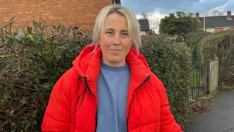 A woman with light blonde hair tied back, wearing a blue sweater and a bright red puffer jacket with black accents
