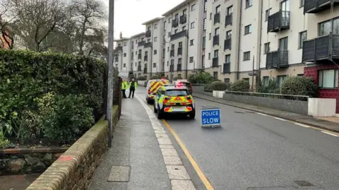 Police cars and a POLICE SLOW sign blocks a main road surrounded by blocks of flats in St Helier.