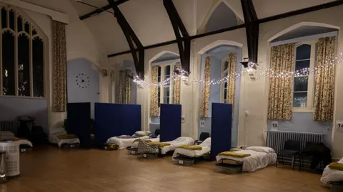 HopeWorks A large room, with blue painted walls, with patterned curtains at each window, which are all open. Fairy lights are strung across the room, and a number of camp beds are made up, with blue panels dividing them. The floor is wooden and a clock is painted on one wall. Each bed has a number and black chairs are placed around the room. 
