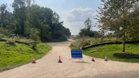 A blue police sign and orange cones blocking the gravel path up to Ham Hill. Either side of the path is surrounded by trees and grass.