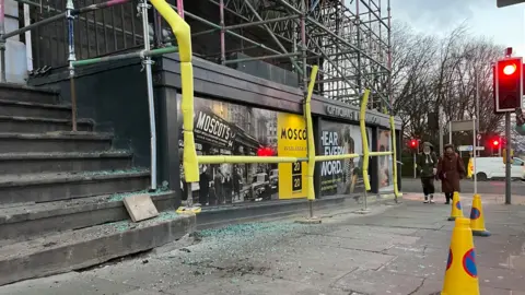 Smashed glass all over the pavement beneath scaffolding. There are yellow cones in the foreground.