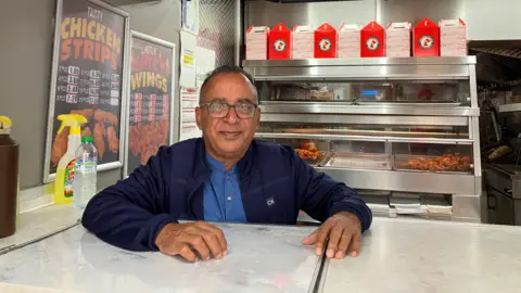 JOANNA TAYLOR/BBC Mohammed Farooq wearing black glasses, a navy jacket and lighter blue shirt is smiling at the camera and resting his hands on a white counter. Behind him are silver counters with fried chicken laid out, red takeaway boxes and posters with price lists.