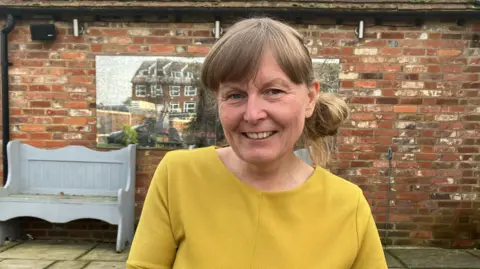 BBC/Amy Holmes Catherine Bunting with light brown hair, tied back, with a fringe. She is wearing a mustard yellow tunic and is sat down in a pub beer garden.