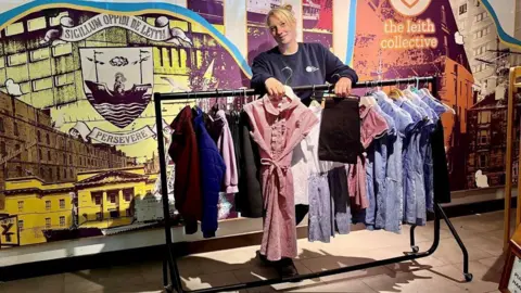 The Leith Collective founder, Sara Thomson, is standing at a rail of secondhand uniforms at one of her stores. She has blonde hair and is wearing a blue jumper. she is holding a pink dress on a hanger.