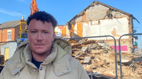 BBC Reece Thomas wearing a hooded coat looking at the camera in front of a demolished building on Saffron Lane in Leicester