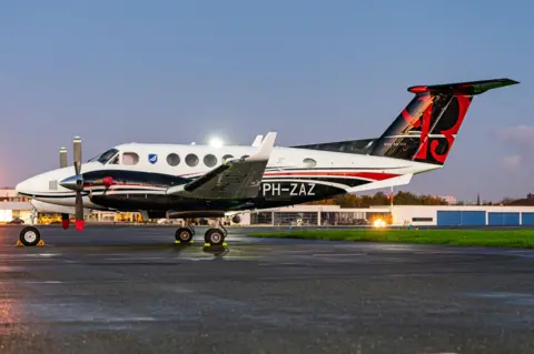 Airteamimages A white and black light aircraft sits on a runway of a small airport with grass and buildings seen behind it