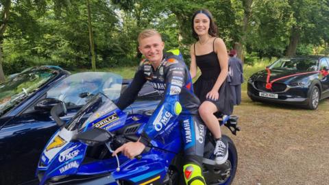 Ryan Vickers with short blond hair and wearing his motorcycle outfit sitting on a blue superbike. Scarlett Alexander, wearing a black dress, is riding on the back.