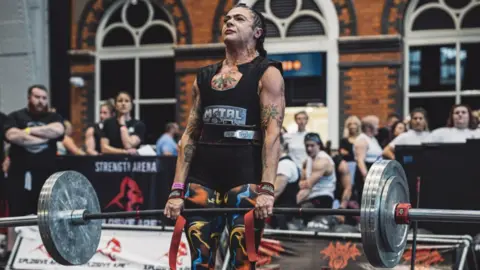 Rosie Abercrombie Rosie is holding a weightlifting bar, grimacing as she keeps in in position during a trial. She has a vest top and tights on with tattoos on her arms and people are standing in the background watching her.
