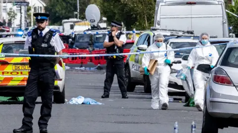 Police and forensics on Hart Street Southport, Merseyside, after stabbings of children at a dance class. There is police tape cordoning off the road, several cars and two people in white suits 