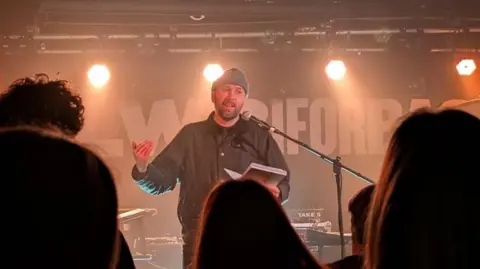 Hayley Lau Matt performing poetry on stage at Clwb Ifor Bach in Cardiff. He stands behind a black microphone and holds his book in his right hand. He wears a black jacket and a grey beanie hat.