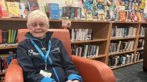 The image shows an elderly woman in a dark blue volunteers jacket sitting in a brown leather armchair in the public library at the University Hospital in Coventry.
