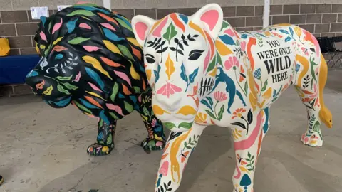 There are two large sculptures of a lion and lioness. The lion is painted black and the lioness is painted white and both have brightly coloured designs and the words You Were Once Wild Here