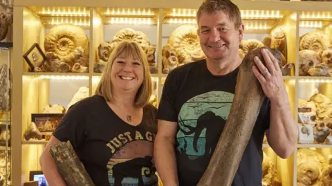 David Vintiner Sally and Neville Hollingworth stand next to each other holding a large mammoth bone each. They stand in front of a brightly-lit cabinet which displays a selection of fossils.