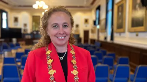 A woman with a red blazer and a black t-shirt underneath. She is wearing a large ceremonial gold chain around her neck. Behind here there rows of blue chairs and big gold-framed paintings on the walls.