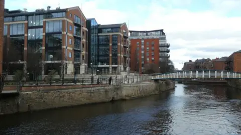 The River Aire in Leeds city centre, with the David Oluwale Bridge in the distance. Flats and offices are on the other side of the water. The sky is blue with some cloud cover. 